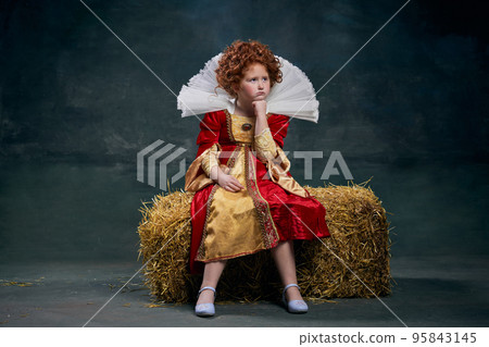Portrait of little red-headed girl, child in costume of royal person on hay isolated over dark green background. Offended face Portrait of little red-headed girl, child in costume of royal person on hay isolated over dark green background. Offended face 95843145