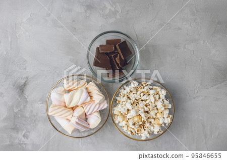 Popcorn, colorful pastel marshmallow, pieces of broken milk chocolate bar in glass bowls on gray background. Ingredients for cooking dessert and unhealthy sweets concept. Top view, copy space Popcorn, colorful pastel marshmallow, pieces of broken milk chocolate bar in glass bowls on gray background. Ingredients for cooking dessert and unhealthy sweets concept. Top view, copy space 95846655