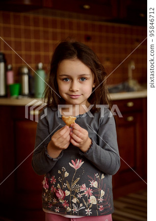 Beautiful Caucasian little girl holding a gingerbread cookie, standing in the home kitchen, cutely smiling looking at the camera Beautiful Caucasian little girl holding a gingerbread cookie, standing in the home kitchen, cutely smiling looking at the camera 95846712