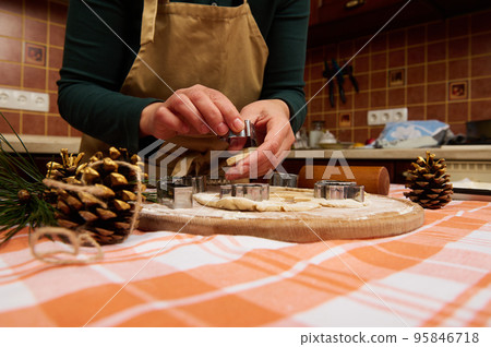 Close-up unrecognizable woman housewife, a confectioner amateur wearing green shirt and beige chef apron, cutting gingerbread dough with cookie cutters on a table. Christmas atmosphere in the kitchen Close-up unrecognizable woman housewife, a confectioner amateur wearing green shirt and beige chef apron, cutting gingerbread dough with cookie cutters on a table. Christmas atmosphere in the kitchen 95846718
