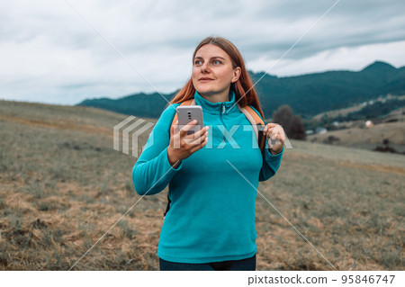 A woman with a backpack stands on the top of a mountain using smartphone and admires the beauty of a mountain valley, a rear view. A woman with a backpack stands on the top of a mountain using smartphone and admires the beauty of a mountain valley, a rear view. 95846747
