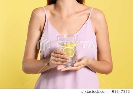 Young woman with glass of lemon water on yellow background, closeup 95847523