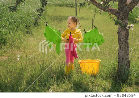 A child hangs Bright colored wet clothes on a clothesline in the garden under a tree on a sunny day. A little girl helps with household chores 95848584