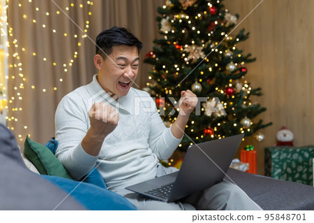 A young Asian man is playing games on a laptop, sitting at home on the couch near the nine-year-old Christmas tree. Happy with success, makes yes with his hands. A young Asian man is playing games on a laptop, sitting at home on the couch near the nine-year-old Christmas tree. Happy with success, makes yes with his hands. 95848701