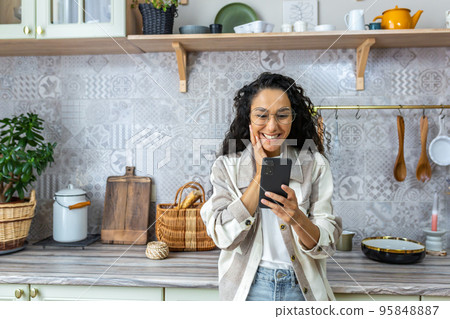 Happy Latin American woman at home reading good online news from smartphone, woman in glasses and curly hair using phone to watch online content at home in kitchen. 95848887