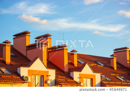 Red rooftop of apartment building against blue sky. Terra cotta tile roof shingles. Newly built modern residential house with chimneys and small windows, clay roofs. Real estate investment, renovation Red rooftop of apartment building against blue sky. Terra cotta tile roof shingles. Newly built modern residential house with chimneys and small windows, clay roofs. Real estate investment, renovation 95849971