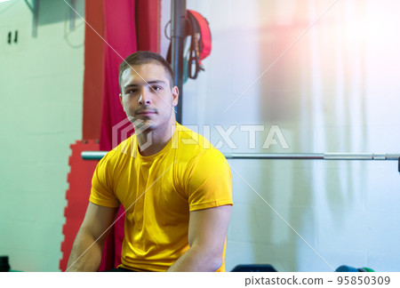 Athletic young man poses seated on the bench press looking at the camera in a break between sets of bench presses. Concept of successful young man, focused on goals. Copy space. 95850309