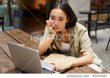 Bored asian girl sitting in cafe and staring at laptop, studying and feeling sad, doing homework Bored asian girl sitting in cafe and staring at laptop, studying and feeling sad, doing homework 95850867