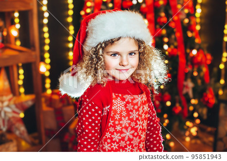 Portraite of little curly girl in Santa hat on blurred lights and christmas tree background.  95851943