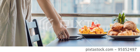 Young woman is laying on a table. Breakfast table with coffee fruit and bread croisant on a balcony against the backdrop of the big city BANNER, long format 95852497