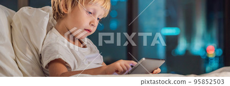 The boy uses the tablet in his bed before going to sleep on a background of a night city. Children and technology concepts BANNER, long format The boy uses the tablet in his bed before going to sleep on a background of a night city. Children and technology concepts BANNER, long format 95852503