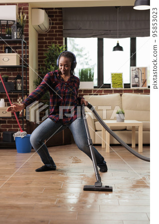 Positive housewife enjoying spring cleaning at home, singing while vacuuming in living room. Listening to music with wireless headphones relaxed looking cheerful, taking care of finishing household Positive housewife enjoying spring cleaning at home, singing while vacuuming in living room. Listening to music with wireless headphones relaxed looking cheerful, taking care of finishing household 95853023