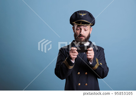Plane pilot in professional uniform playing mobile games on smartphone, holding phone, watching online video. Airlane captain using aviation simulator on telephone, leisure entertainment 95853105