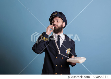 Middle aged pilot talking on landline phone, holding telephone, having conversation. Airlane captain in professional uniform answering call in airport, studio medium shot 95853112