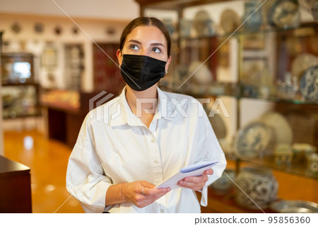 Young girl in mask holding brochure and looking at ceramic exhibits Young girl in mask holding brochure and looking at ceramic exhibits 95856360
