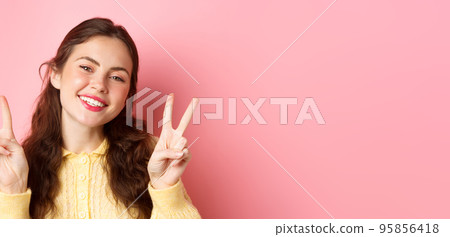 Close up of beautiful young woman showing peace v-sign and smiling happy at camera, wearing bright glam make up, standing against pink background 95856418