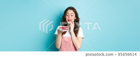 Holidays and celebration. Cute birthday girl looking at tasty cake with temptation to bite it, standing dreamy against blue background Holidays and celebration. Cute birthday girl looking at tasty cake with temptation to bite it, standing dreamy against blue background 95856614