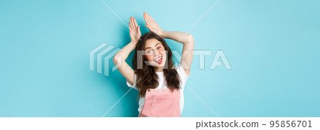 Portrait of beautiful glamour girl celebrating Easter, showing bunny rabbit ears with hands above head, smiling joyful, standing over blue background 95856701