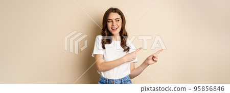 Stylish young caucasian woman smiling, pointing fingers left, showing advertisement, promo offer, standing against beige background 95856846