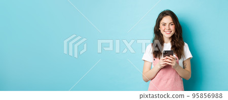 Young cute girl holding mobile phone, smiling and looking at camera. Woman using smartphone app, chatting on social media or shopping online, blue background 95856988