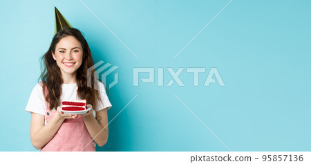 Holidays and celebration. Cheerful birthday girl in party hat holding bday cake and smiling, making wish on lit candle, standing against blue background Holidays and celebration. Cheerful birthday girl in party hat holding bday cake and smiling, making wish on lit candle, standing against blue background 95857136