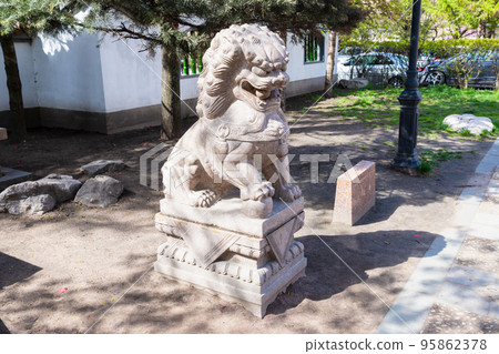 Lion sculpture statue with ball in his mouth and with ball under right paw in Chinese park. Mythical guardian lion. Close-up, side view. Garden of Friendship Lion sculpture statue with ball in his mouth and with ball under right paw in Chinese park. Mythical guardian lion. Close-up, side view. Garden of Friendship 95862378