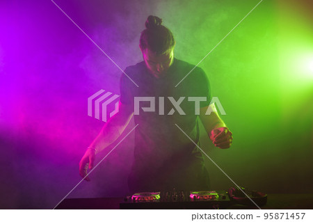 DJ at work mixing sound on her decks at a party or night club with colourful smoke light background DJ at work mixing sound on her decks at a party or night club with colourful smoke light background 95871457