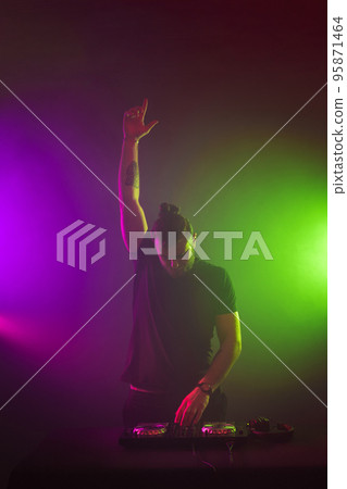 DJ at work mixing sound on her decks at a party or night club with colourful smoke light background DJ at work mixing sound on her decks at a party or night club with colourful smoke light background 95871464