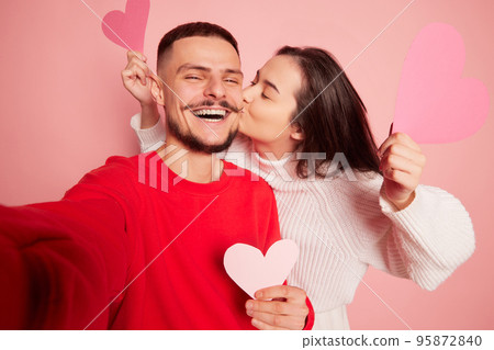 Portrait of lovely young couple, woman kissing happy and smiling man, taking selfie together isolated over pink background Portrait of lovely young couple, woman kissing happy and smiling man, taking selfie together isolated over pink background 95872840