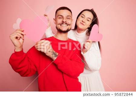 Portrait of lovely young couple, man and woman posing with paper hearts isolated over pink background. Holiday party Portrait of lovely young couple, man and woman posing with paper hearts isolated over pink background. Holiday party 95872844