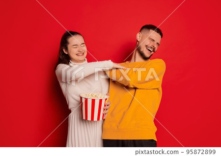 Portrait of happy young couple, man and woman, posing with popcorn isolated over red background. Giggling, playing 95872899