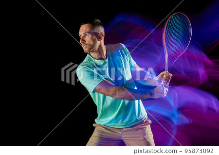 Movements in neon glow. Active male tennis player in sportwear playing tennis isolated over dark background in mixed purple neon light. 95873092