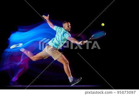Young male tennis player in t-shirt and shorts playing tennis isolated over dark background in mixed purple neon light. Competition, creativity, art, ad 95873097