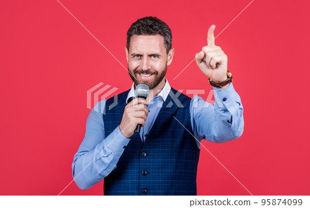 Talk show host. Happy speaker speak into microphone keeping finger raised. Speaker perform speech. Visiting professor or lecturer. Speaking coach. Hosting conference. Voice professional 95874099