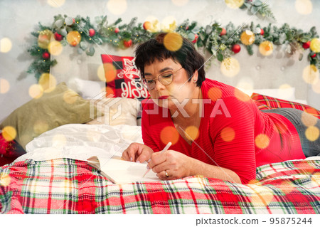 happy elderly woman with pen and notebook making wish list or to do list for new year in bed over christmas tree. xmas holidays concept 95875244