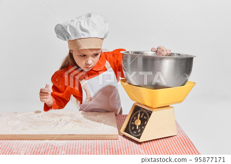 Cute little child, girl in image of chef cooking over grey background. Measuring flour Cute little child, girl in image of chef cooking over grey background. Measuring flour 95877171