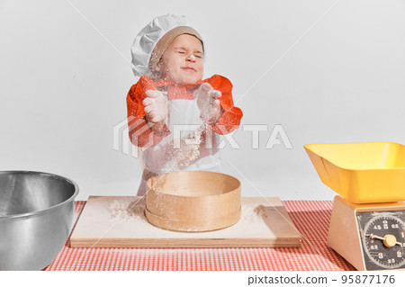 Cute little child, girl in image of chef cooking over grey background. Sifting flour, dirty face Cute little child, girl in image of chef cooking over grey background. Sifting flour, dirty face 95877176