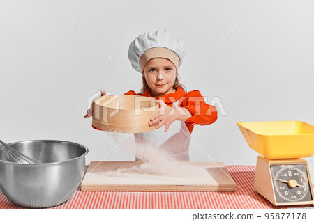 Cute little child, girl in image of chef cooking over grey background. Sifting flour Cute little child, girl in image of chef cooking over grey background. Sifting flour 95877178