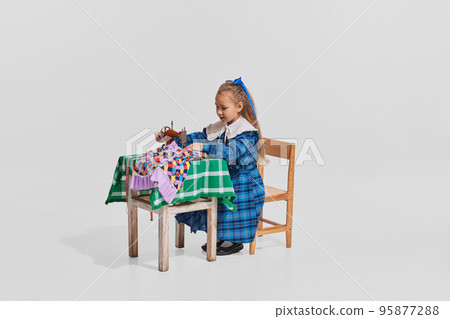 Portrait of pretty little girl in cute blue dress sitting at the table and sewing isolated over grey background Portrait of pretty little girl in cute blue dress sitting at the table and sewing isolated over grey background 95877288