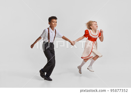 Portrait of beautiful little children, boy and girl in vintage clothes holding hands and running isolated over grey background 95877352