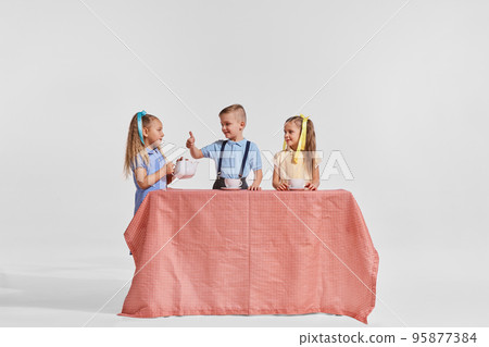 Portrait of three children, boy and girls sitting at the table and drinking tea over grey background. Breakfast time 95877384