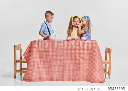 Portrait of three children, boy and girls sitting at the table over grey background. Girls whispering secrets 95877395