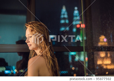 Beautiful woman wipes her hair after a shower on the background of a window with a panoramic view of the night city 95877502