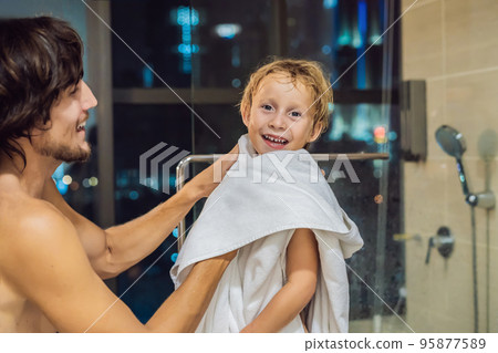 Dad wipes his son with a towel after a shower in the evening before going to sleep on the background of a window with a panoramic view of the city Dad wipes his son with a towel after a shower in the evening before going to sleep on the background of a window with a panoramic view of the city 95877589
