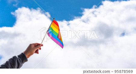 A hand in a hoodie holds an LGBT flag in the fingers in the wind on a cloudy background 95878569