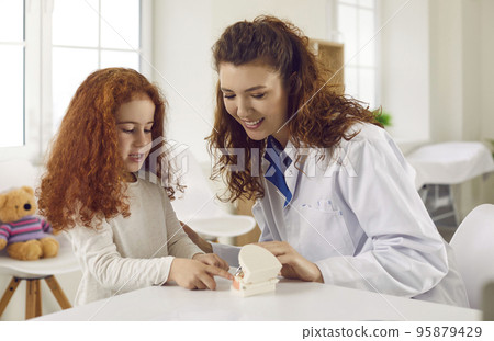 Female dentist tells little girl about oral hygiene by showing her artificial jaw with teeth. Female dentist tells little girl about oral hygiene by showing her artificial jaw with teeth. 95879429