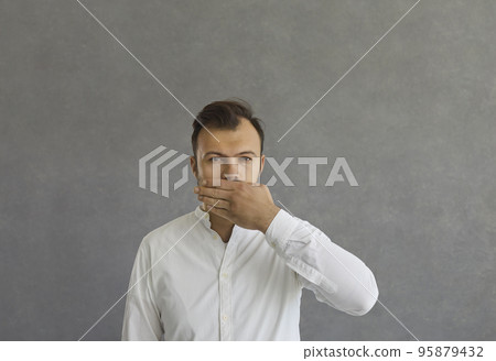 Handsome serious man closes his mouth with his hand while standing on a gray concrete background. 95879432