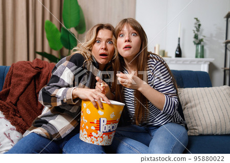 Mother and daughter having fun spending leisure time together at home, watching scary movie on TV and eating popcorn 95880022