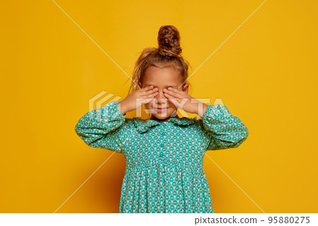 Partial image of serious little girl closed eyes with his hands. Portrait of caucasian female child wearing color dress. Childhood concept. Isolated on yellow studio background. Copy space 95880275