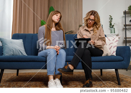 Mature female psychologist listens attentively to young woman and writes in clipboard while sitting on couch 95880482